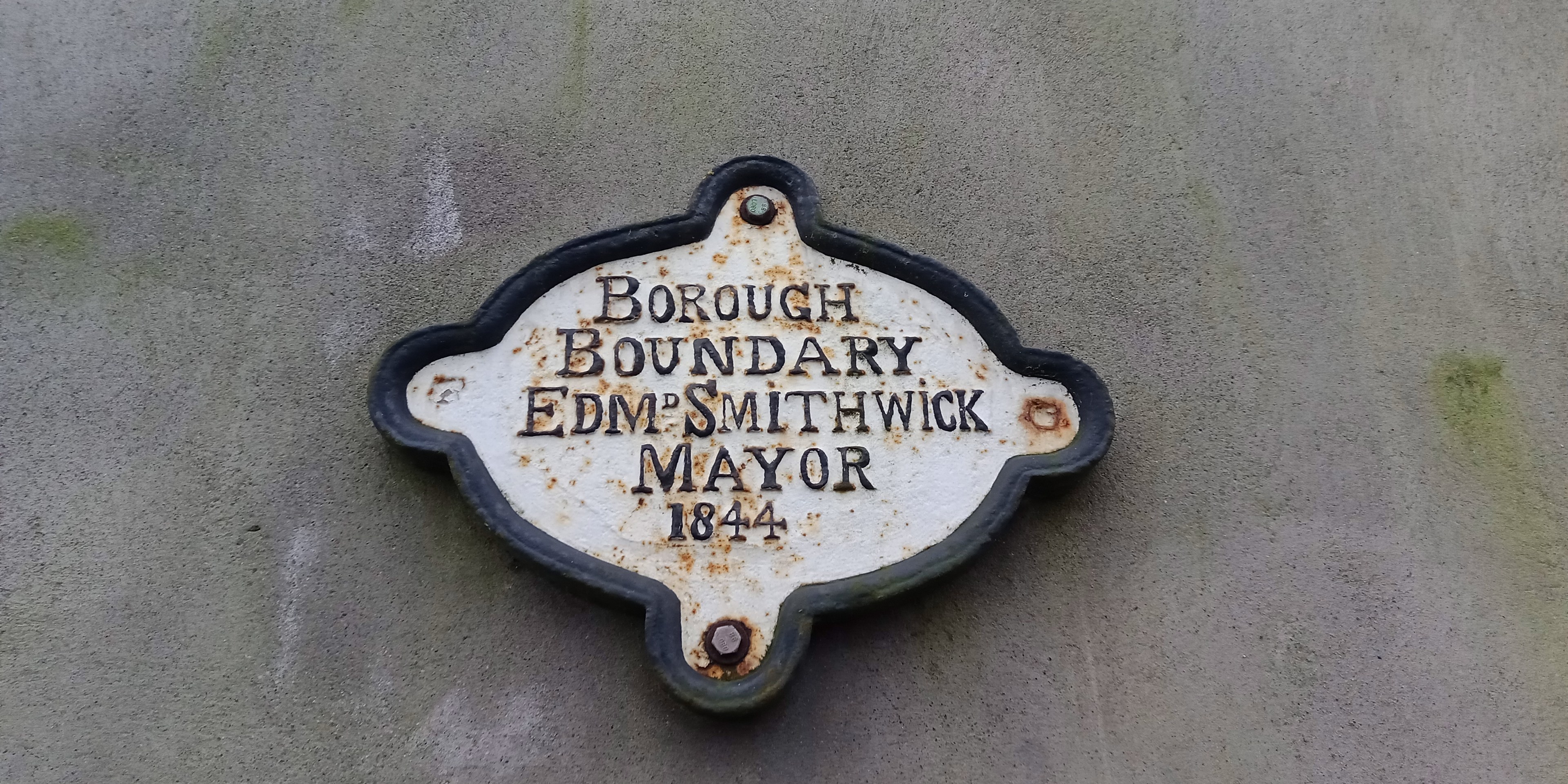 Metallschild an einer Gebäudeseite mit schwarzem Hintergrund und weißer fettgedruckter Schrift, die "Borough Boundary Edm Smithwick Mayor" lautet.