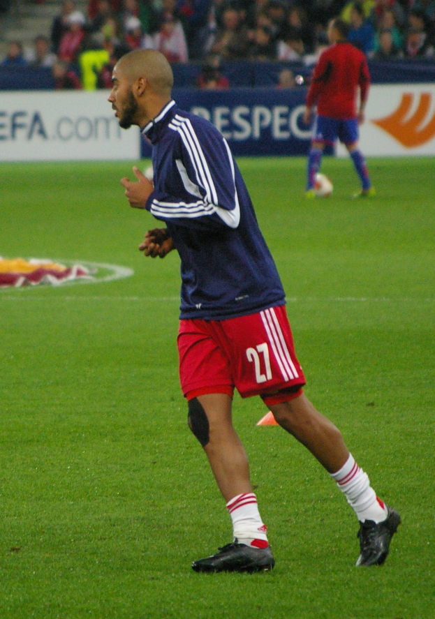 Ein Mann in einem blauen Hemd und roten Shorts läuft auf einem Fußballfeld, umgeben von sitzenden Zuschauern und einigen Werbetafeln, trägt Schuhe.