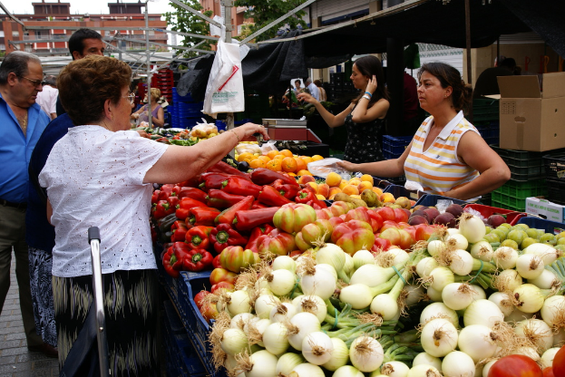 Eine Gruppe von Menschen, die sich draußen in der Nähe von Gebäuden und Bäumen aufhalten, mit Gemüse in Plastikbehältern und einer Kartonschachtel in der Nähe.