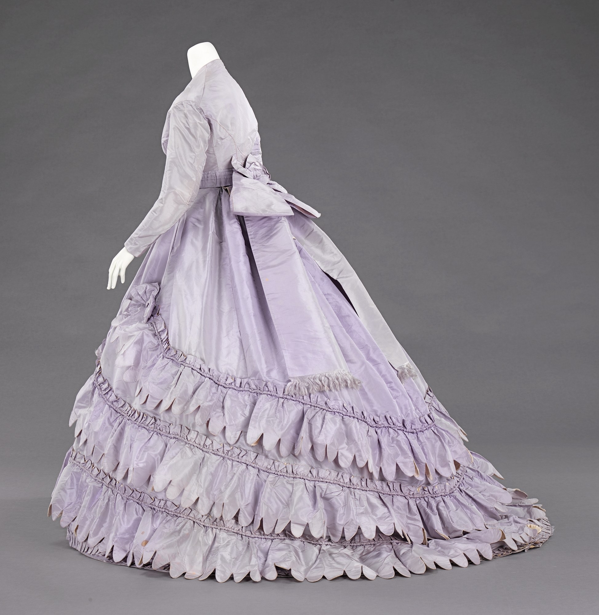 Eine Schaufensterpuppe in einem lila Rüschenkleid mit engem Oberteil und vollem, bodenlangen Rock vor einem grauen Hintergrund.