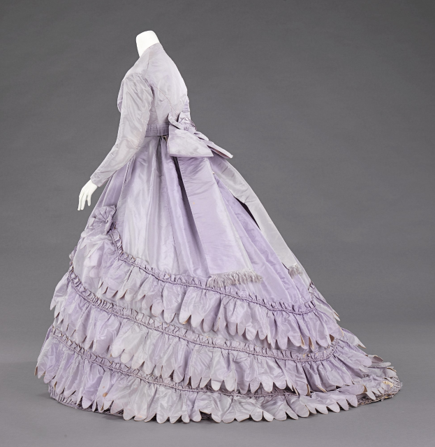 Eine Schaufensterpuppe in einem lila Rüschenkleid mit engem Oberteil und vollem, bodenlangen Rock vor einem grauen Hintergrund.