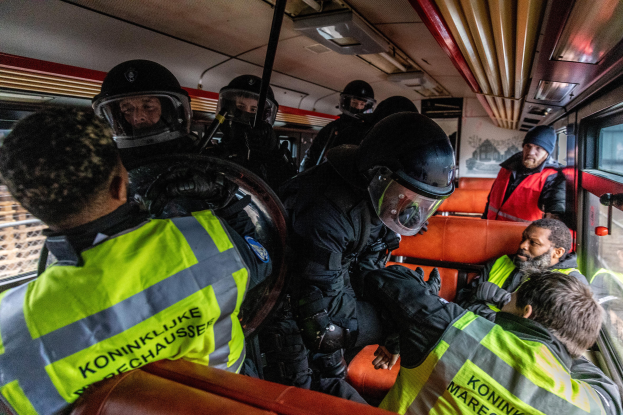 Eine Gruppe von Polizisten in Einsatzausrüstung auf einem Bus, mit einer Person auf dem Sitz in der Mitte und einem Plakat im Hintergrund.
