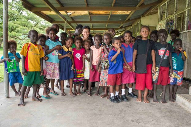 Eine Gruppe von Kindern steht vor einem Gebäude, mit einer Frau in der Mitte, Treppen auf der rechten Seite, einem Pfahl auf der linken Seite und üppiger Vegetation im Hintergrund.