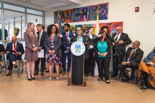 Eine Gruppe von Menschen steht und sitzt in einem Raum mit einem Podium, einem Mikrofon und einer Tafel mit der Aufschrift "Sancho Stadtrat tritt dem Stadtrat bei", mit Flaggen, einer Tafel, einem Feuerlöscher und Deckenleuchten an der Rückwand.