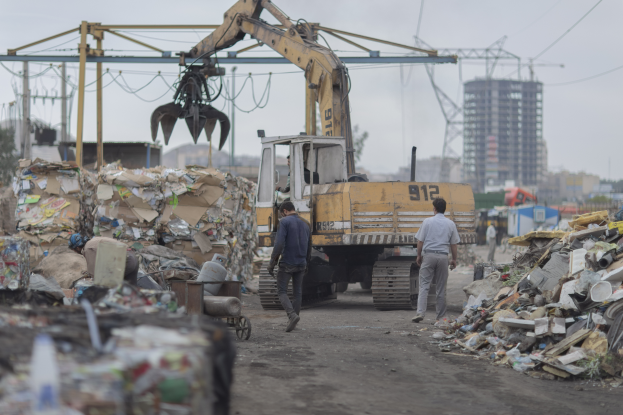 Eine Gruppe von Männern, die einen Schotterweg neben einem Haufen Müll entlanggehen, mit einem Bagger im Hintergrund, Gebäuden, Bäumen und anderen Objekten in der Umgebung.