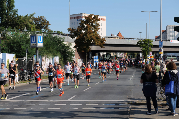 Gruppe von Menschen, die bei einem Marathon auf einer baumgesäumten Straße mit Laternen, Schildern, Fahrrädern, einem Zaun, Gras, einer Brücke, Gebäuden und einem klaren blauen Himmel laufen.