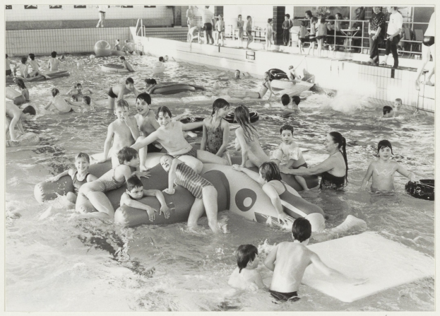 Schwarzes und weißes Bild von Kindern, die in einem Swimmingpool spielen, einige auf aufblasbaren Gegenständen, andere stehend, mit einer Wand und einem Geländer im Hintergrund.