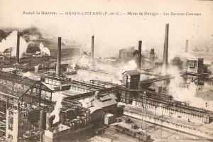 Ein altes Schwarz-Weiß-Foto einer Fabrik mit Rauch, der aus ihren Schornsteinen aufsteigt, umgeben von Gebäuden, Masten und Eisenbahnschienen, mit dem Text "Avant la Guerre - Hénin-Lietard - Mines de Douges - Les Services Centraux" oben und unten.