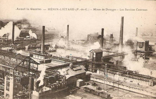 Ein altes Schwarz-Weiß-Foto einer Fabrik mit Rauch, der aus ihren Schornsteinen aufsteigt, umgeben von Gebäuden, Masten und Eisenbahnschienen, mit dem Text "Avant la Guerre - Hénin-Lietard - Mines de Douges - Les Services Centraux" oben und unten.