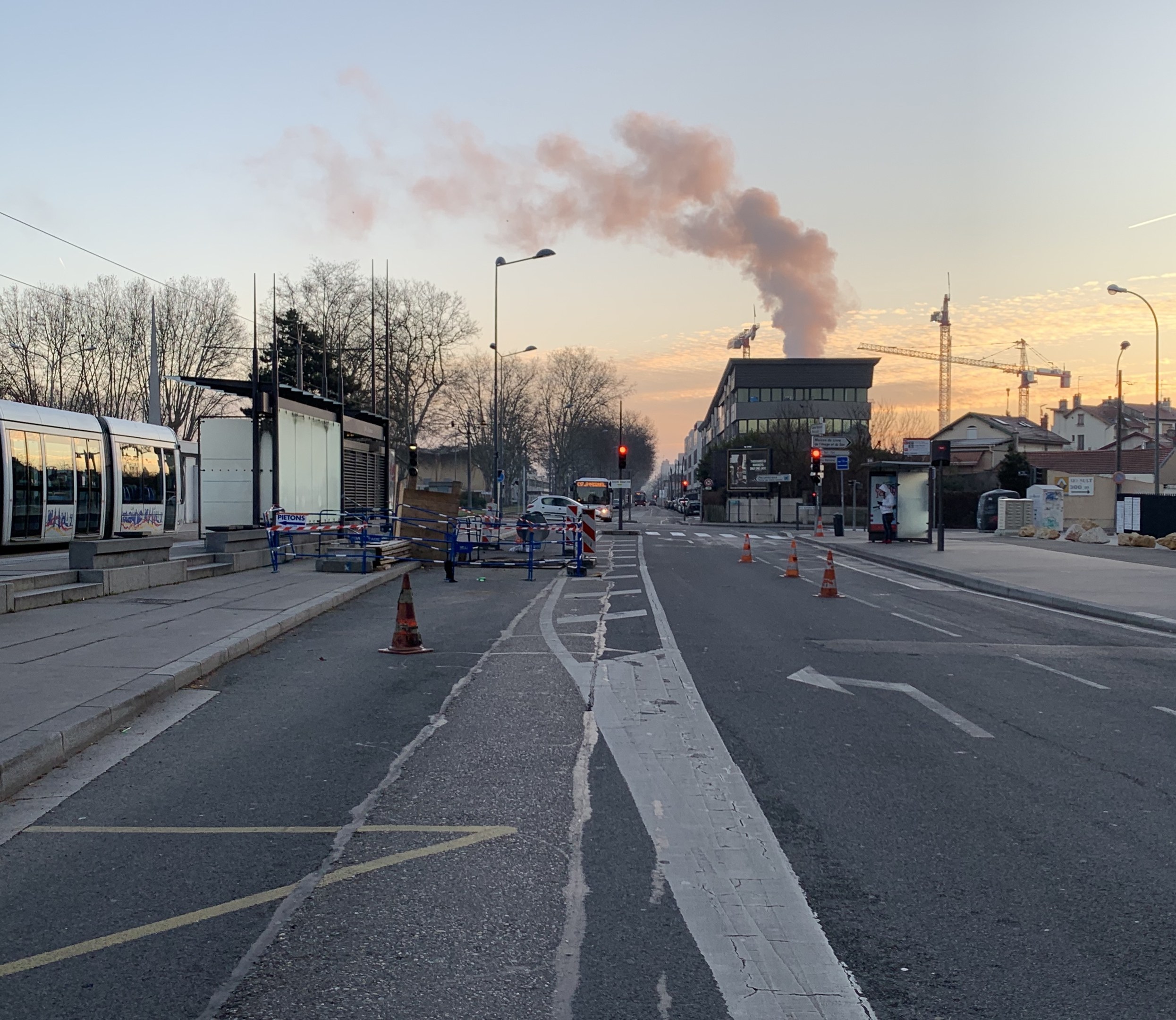 Eine Stadtstraße mit Absperrbaken, einem Bus, Laternen, Bäumen, Gebäuden, Kränen und Rauch, der aus einem entfernten Gebäude aufsteigt.