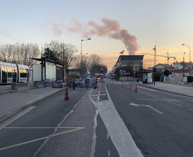 Eine Stadtstraße mit Absperrbaken, einem Bus, Laternen, Bäumen, Gebäuden, Kränen und Rauch, der aus einem entfernten Gebäude aufsteigt.
