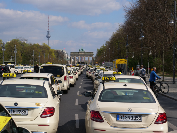 Eine lange Reihe von Taxis, die entlang einer belebten Straße in Berlin, Deutschland, geparkt sind, mit Fahrradfahrern und Fußgängern auf dem Gehweg, flankiert von Bäumen und Laternenmasten und Gebäuden, einem Bogen und einem Turm im Hintergrund.