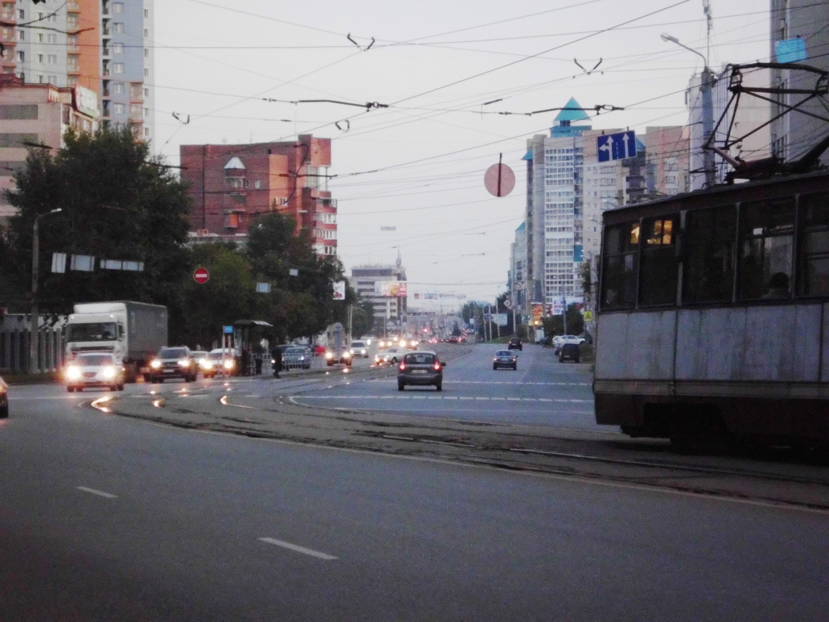 Stadtstraße mit starkem Verkehr einschließlich eines Busses, Lastwagens und einer Straßenlaterne, umgeben von Schildern, Bäumen, Pfählen, Drähten, Gebäuden und einem klaren blauen Himmel.