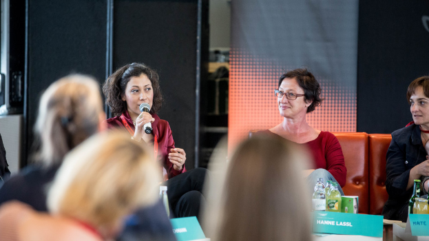 Eine Gruppe von Frauen sitzt in der Mitte, wobei eine auf der linken Seite ein rotes Kleid trägt und ein Mikrofon hält, und ein rollbares Banner und ein schwarzes Tuch im Hintergrund.