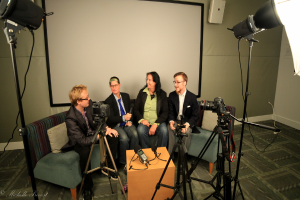 Eine Gruppe von drei Personen sitzt auf einem Sofa und gibt ein Interview, wobei eine Person in einem schwarzen Mantel und Hose links, eine Person in einem Mantel und blauer Krawatte mit einem Mikrofon in der Mitte und eine strahlende Person rechts zu sehen ist. Ein Mikrofonrekorder befindet sich auf dem mittigen Tisch, Kameras sind auf der rechten und linken Seite und große Lichtstative sind vorhanden, mit einem Projektionsschirm im Hintergrund.