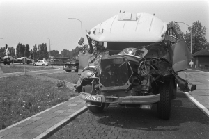 Ein Schwarz-Weiß-Foto eines schwer beschädigten Autos am Straßenrand mit eingedellter Haube und zersplitterten Fenstern sowie anderen Fahrzeugen, Laternenpfählen, Bäumen und einem Haus im Hintergrund.