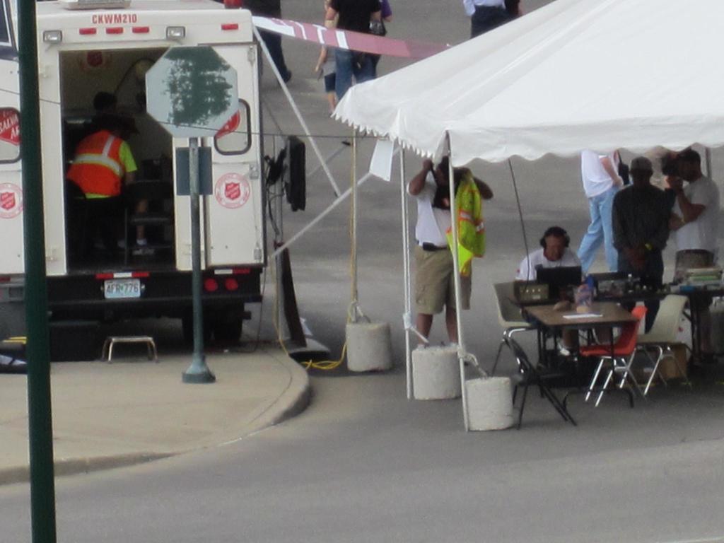 Ein Krankenwagen steht auf der linken Seite einer Straße, während auf der rechten Seite ein Zelt eine Gruppe von Menschen schützt, bei der ein Tisch und ein Stuhl in der Nähe stehen.