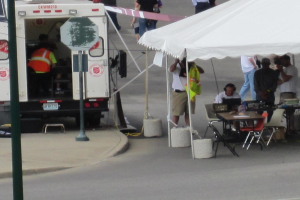 Ein Krankenwagen steht auf der linken Seite einer Straße, während auf der rechten Seite ein Zelt eine Gruppe von Menschen schützt, bei der ein Tisch und ein Stuhl in der Nähe stehen.