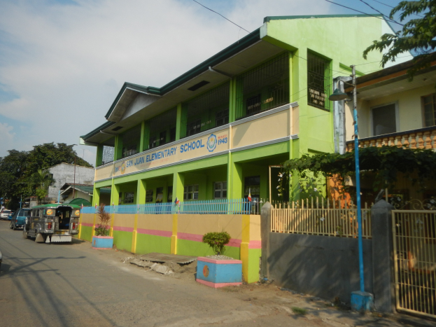 Ein Schulgebäude mit grüner und gelber Fassade, einem Zaun, einem Tor, einer Wolke und einer Straße mit Fahrzeugen, Topfpflanzen, einer Straßenlaterne, Bäumen und Drähten.