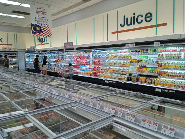 Ein gut sortierter Saftladen mit Regalen voller Getränke, frischem Obst und anderen Artikeln, einigen Kunden, die durch die Gänge schlendern, und einer Deckenleuchte, die den Raum erhellt.