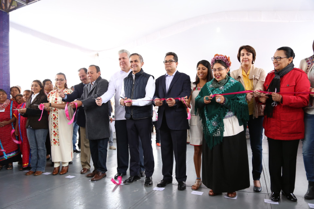 Eine Gruppe von Menschen, die eine Schnur schneiden, bei einer Zeremonie, die lächeln, mit einer Säule links, verstreuten Papieren auf dem Boden und einem weißen Tuch im Hintergrund.